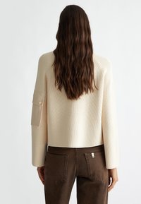Maglione a coste color crema con un design accorciato e una tasca laterale con bottone. Indossato sopra pantaloni marroni con tasche posteriori.