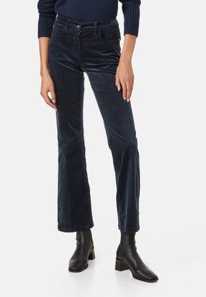 Vrouw draagt marineblauwe corduroy flare broek met zwarte enkellaarzen en een donker marineblauwe trui, staand tegen een witte achtergrond.