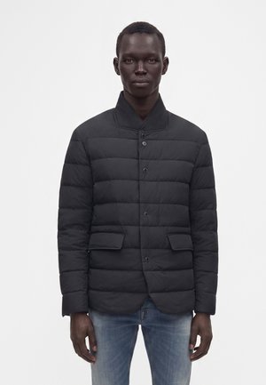 Mann trägt eine schwarze gesteppte Jacke mit Knöpfen und Klappentaschen sowie blaue Jeans, steht vor einem einfarbigen hellen Hintergrund.