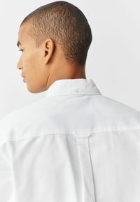 Camisa blanca de botonadura con un collar estructurado, que presenta una abertura en la parte posterior y una textura de tela suave. Tono neutro con detalles mínimos.