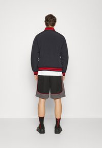 Marineblaues Bomberjackett mit rot gestreiftem Kragen, schwarze Shorts mit grauen Akzenten und schwarze Sneakers. Ein sportliches, lässiges Outfit.