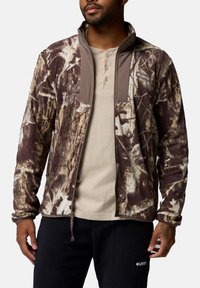 Tarnfleecjacke in Braun- und Beigetönen mit hohem Kragen, Reißverschluss vorne und zwei Taschen. Mit einem Logo auf der Brust.
