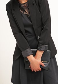 Czarna marynarka o dopasowanym fasonie i zapięciu na guziki, założona na cz dress z koronkowymi detalami, połączona z czarnym kopertówką.
