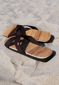 Brown fabric sandals with wooden textured footbeds, featuring cross straps and a black rubber sole, resting on sandy surface.