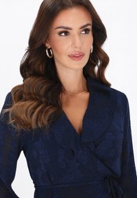 Tengerészkék ruha virágos textúrával, fodros gallérral, átívelő kialakítással és megkötős derékkal. Az arany karika fülbevalók teszik teljessé a megjelenést.