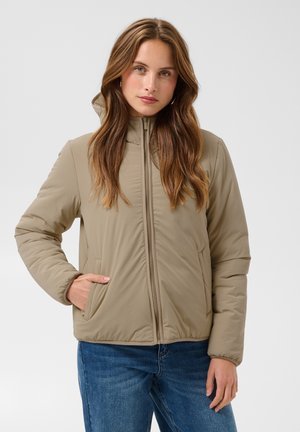 Femme aux longs cheveux bruns portant une veste beige zippée et un jean bleu, debout avec une main dans la poche devant un fond uni.