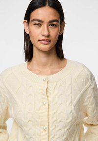Cardigan lavorato a maglia bianco sporco con motivo a trecce, scollatura rotonda e bottoni perlescenti, rifinito con bordi a costine e dettagli testurizzati.