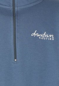 Pullover in pile blu con zip a quarto. Presenta una scritta ricamata bianca che legge "downtown" e "Καoticο" sul petto. Texture liscia.