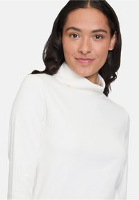 Weißes, geripptes Rollkragenoberteil mit langen Ärmeln, das eine weiche Textur und eine figurbetonte Passform aufweist, getragen von einem Modell mit dunklem, welligem Haar.