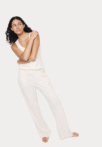 Weißer ärmelloser Jumpsuit mit entspannter Passform, vertikaler gerippter Textur und weitem Bein. Model steht barfuß mit verschränkten Armen.