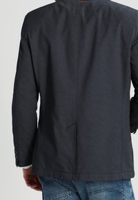 Marineblazer aus strukturiertem Stoff, mit einem Schlitz hinten und Drei-Knopf-Manschetten, getragen über Jeans.