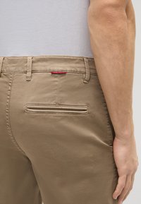 Pantalones de algodón beige con una textura suave, que cuentan con un bolsillo trasero y detalle de etiqueta roja cerca de la cintura. Diseño liso, corte ajustado.