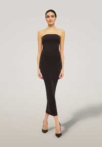 Donna in piedi che indossa un vestito midi nero aderente senza spalline e tacchi alti neri contro uno sfondo chiaro e semplice.