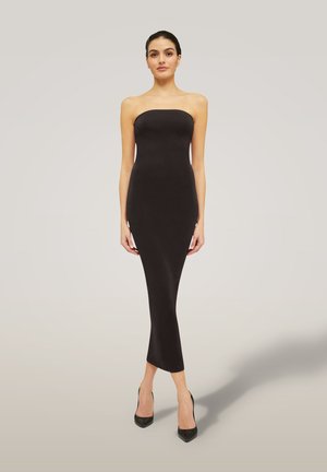 Donna in piedi che indossa un vestito midi nero aderente senza spalline e tacchi alti neri contro uno sfondo chiaro e semplice.