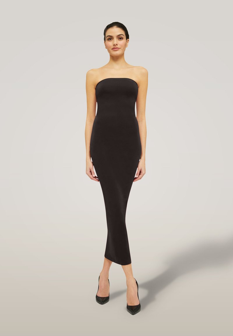 Donna in piedi che indossa un vestito midi nero aderente senza spalline e tacchi alti neri contro uno sfondo chiaro e semplice.