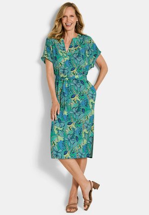 Femme souriante portant une robe bleu sarcelle à fleurs, à manches courtes et longueur genou, avec une ceinture à la taille, debout avec une main dans la poche et les jambes croisées.