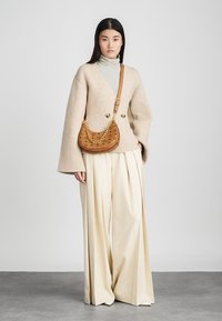 Pull en laine beige à manches évasées, associé à un pantalon crème plissé. Le mannequin porte un sac à main marron, courbé, avec une bandoulière en chaîne dorée.