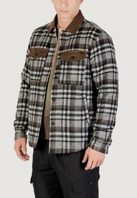 Camicia in flanella a quadri nei toni di marrone, nero e bianco. Presenta dettagli in velluto a coste marrone, due tasche frontali e chiusura a bottoni.