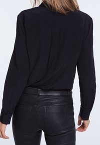 Personne portant une chemise noire à manches longues rentrée dans un pantalon en cuir noir taille haute, vue de dos sur un fond uni.