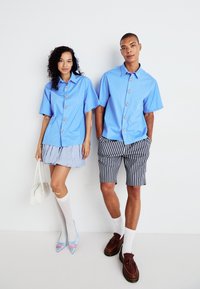 Lyseblå kortermede skjorter med blomstermotiverte knapper, sammen med et rutete miniskjørt og stripete shorts. Tilbehøret inkluderer en hvit håndveske og sokker.