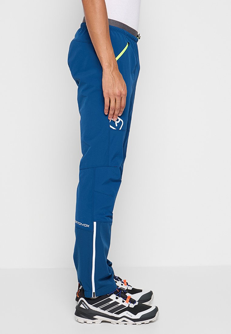 Vue de côté d'une personne portant un pantalon de plein air bleu avec des chevilles fuselées zippées, un petit logo sur la cuisse et des chaussures de randonnée noir-blanc avec des accents orange.