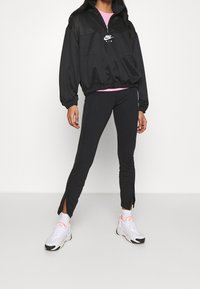Kvinna iklädd svart Nike Air sweatshirt, svarta leggings med ankelsnitt, vita strumpor och vita sneakers med rosa detaljer, stående mot vit bakgrund.