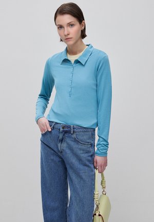 Junge Person mit kurzen Haaren, die ein blaues langärmeliges Polo und lockere blaue Jeans trägt und eine blassgelbe Handtasche an der Seite hält.