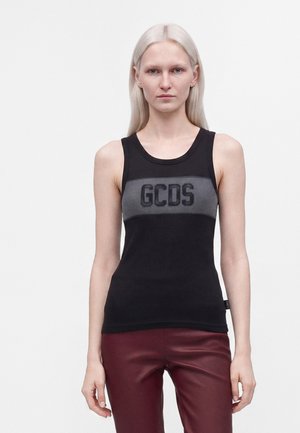 Femme mince aux cheveux blonds platine et lisses porte un haut noir sans manches avec un logo gris "GCDS" et un pantalon rouge foncé sur fond blanc.