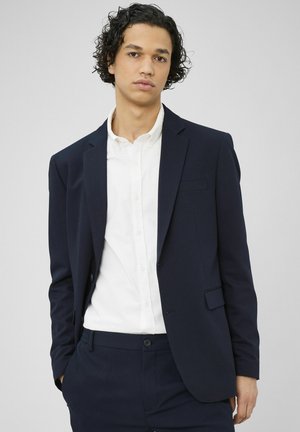 Jeune homme aux cheveux bouclés portant un costume bleu marine composé d’une veste et d’un pantalon, avec une chemise blanche boutonnée, sur un fond uni.