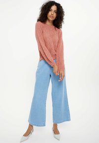 Gehaakte roze trui met een gestructureerd patroon, gecombineerd met lichtblauwe wijde jeans en heldergrijze hakken.