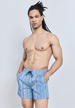 Homme torse nu avec des dreadlocks portant un short de bain rayé bleu, debout les mains dans les poches sur un fond gris uni.