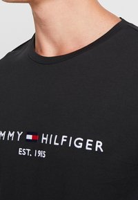 Schwarzes Baumwoll-T-Shirt mit Rundhalsausschnitt, mit weißem gesticktem Text "TOMMY HILFIGER EST. 1985" und einem kleinen Logo in Rot und Blau.