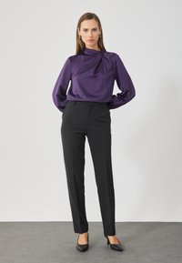 Bluză din satin violet cu detalii drapate, purtată cu pantaloni negri croiți. Croială slim și textură netedă, completate de tocuri negre.