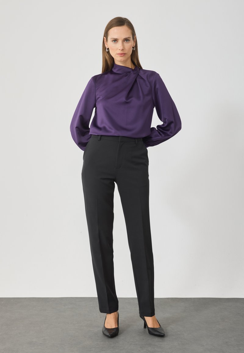 Bluză din satin violet cu detalii drapate, purtată cu pantaloni negri croiți. Croială slim și textură netedă, completate de tocuri negre.
