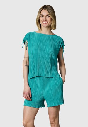 Femme souriante portant un top plissé bleu sarcelle à manches courtes avec des liens et un short plissé assorti, debout devant un fond clair uni.