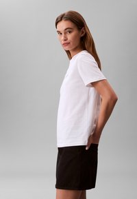 Fehér, rövid ujjú pamut póló, kerek nyakkivágással és karcsúsított szabással. Sötét rövidnadrággal viselve, amely oldalzsebbel rendelkezik.