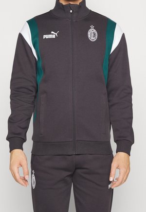 Man draagt een zwart Puma AC Milan trainingsjack met groene en witte accenten op de schouders, dichtgeritst, en bijpassende zwarte broek.
