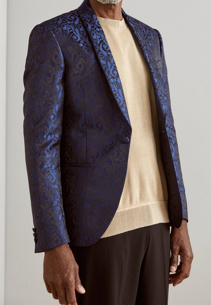 Blazer da uomo in tessuto jacquard blu scuro con motivo paisley, chiusura con un bottone e sottostrato in maglia beige a contrasto.