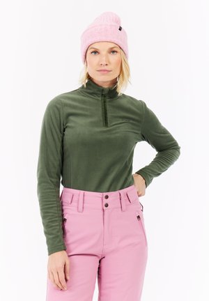Vrouw in een roze gebreide beanie, olijfgroene fleece top met lange mouwen en hooggewaagde roze broek met ritszakken, staande tegen een witte achtergrond.