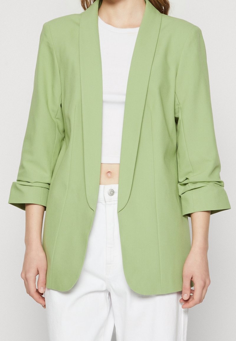 Ljusgrön kavaj med skräddarsydd passform, slagkrage och trekvartslånga ärmar, matchad med en vit crop top och vita byxor.