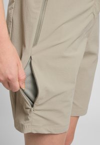 Beige Shorts mit einer Reißverschlusstasche, die ein graues Netzfutter zeigt. Der Stoff ist leicht und glatt, mit einem eleganten Design und klaren Linien.