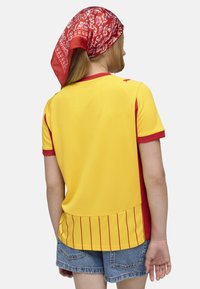 En person iført en gul og rød sportstrøje med et rødt mønstret tørklæde, set bagfra, stående i afslappede denimshorts.