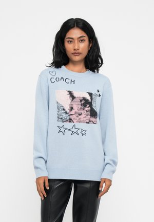 Un pull en tricot bleu clair avec une image graphique, des accents noirs et roses, ainsi que des étoiles décoratives et du texte sur le devant.