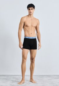 Boxer shorts neri con elastico blu etichettato "HUGO." Il materiale sembra liscio; il design è aderente e di lunghezza a metà coscia.
