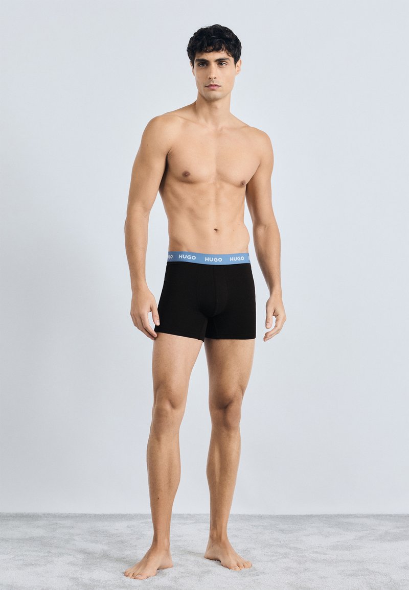 Boxer shorts neri con elastico blu etichettato "HUGO." Il materiale sembra liscio; il design è aderente e di lunghezza a metà coscia.