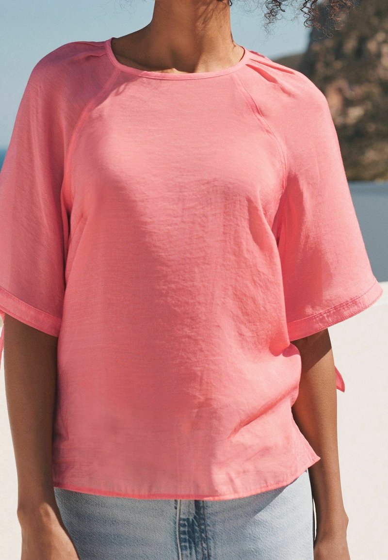 Femme portant un haut corail rose ample à manches courtes et un pantalon en jean bleu clair, debout à l'extérieur avec un arrière-plan naturel flou.