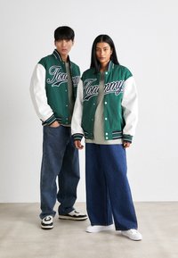 Casacos varsity verdes e brancos com letras "Tommy", combinados com jeans azuis largos. Ténis brancos e pretos, t-shirts cinzentas por baixo.