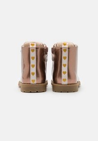 Par de botas metálicas en color rosa oro para toddlers con suelas beige y tiras traseras blancas que presentan un patrón de corazones amarillos, mostradas desde la parte trasera.