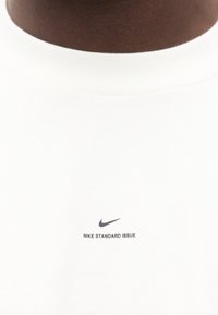 Κοντινό πλάνο σε λευκό μπλουζάκι με στρογγυλή λαιμόκοψη, με μικρό μαύρο λογότυπο Nike και το κείμενο «NIKE STANDARD ISSUE» κάτω από τη λαιμόκοψη.