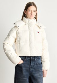 Kurtka puchowa o skróconym kroju w kolorze off-white, z wysokim kołnierzem, zatrzaskami i subtelnym logo. Połączona z ciemnoniebieskimi dżinsami.